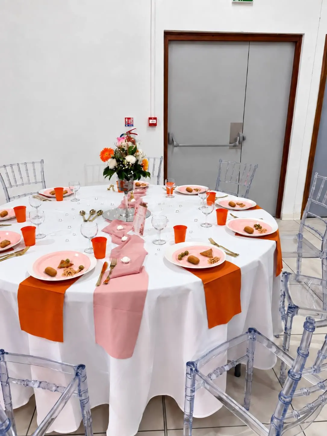 Table ronde dressée en rose et orange, bouquet de fleurs et chaises transparentes